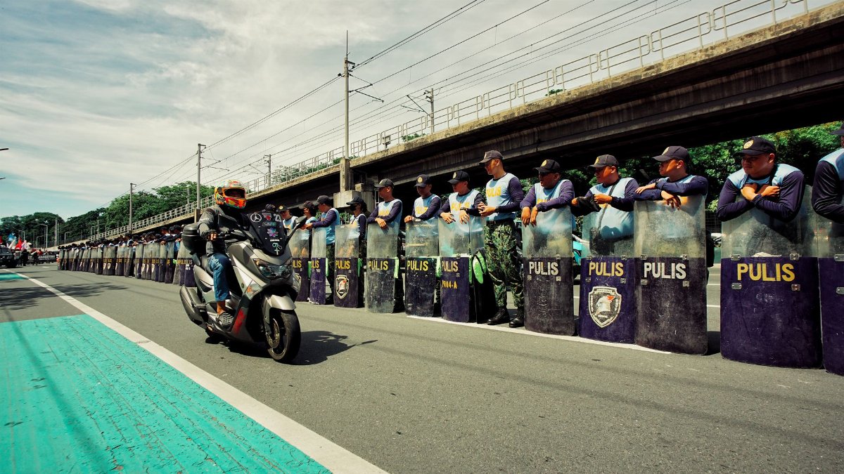 A motorbike passes a line of policemen with shields on an urban street.