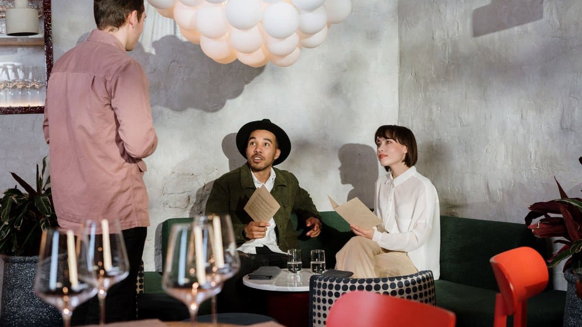 A couple reviews a menu at a chic restaurant, engaging with a waiter.