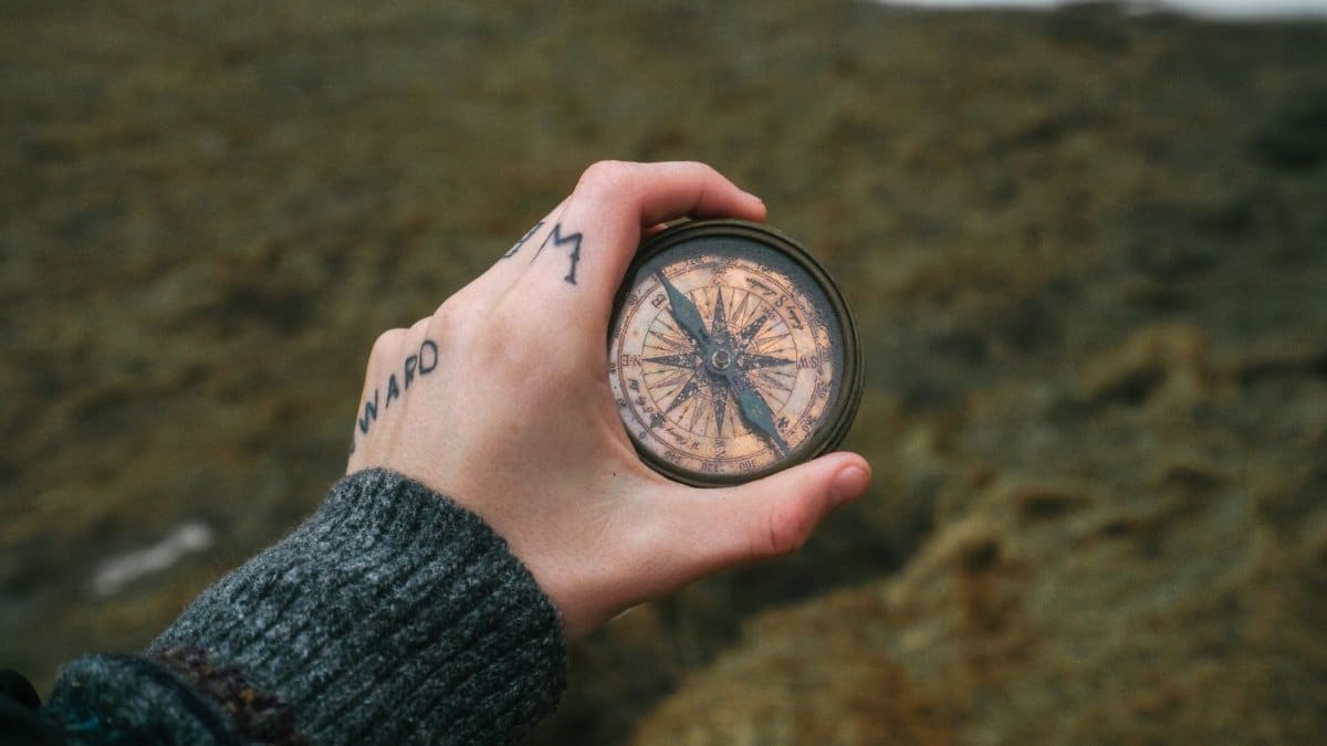 A hand with tattoos holding an antique compass, symbolizing exploration and navigation in nature.