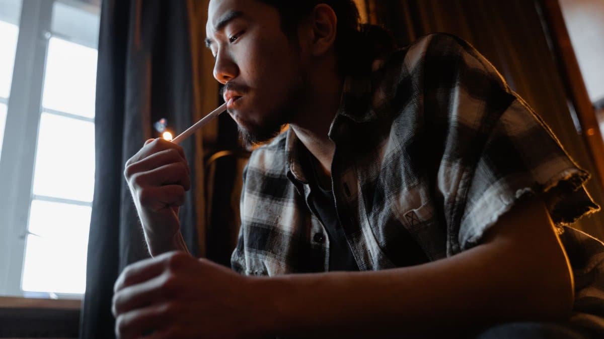 A young man in a plaid shirt lighting a cigarette indoors, illustrating a smoking habit.