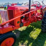 A classic red tractor on display at a rural farm fair under a bright blue sky.