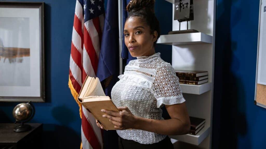 Professional woman holding a book poses confidently in an office setting with flags in the background.