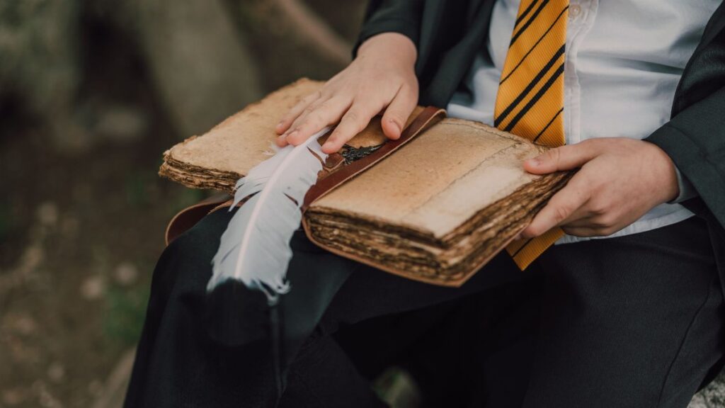 A child in a wizard costume reading an ancient spell book outdoors using a feather quill.