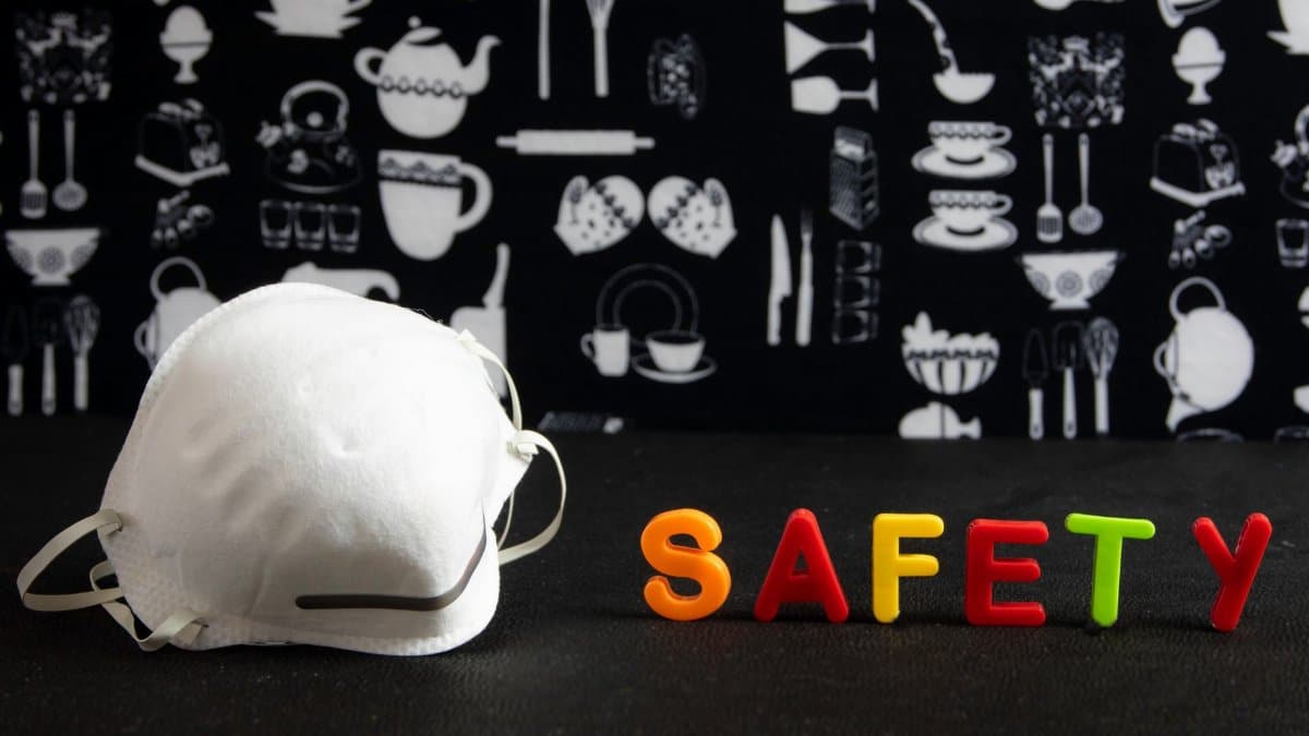 Face mask next to colorful 'SAFETY' letters against a kitchen-themed background.