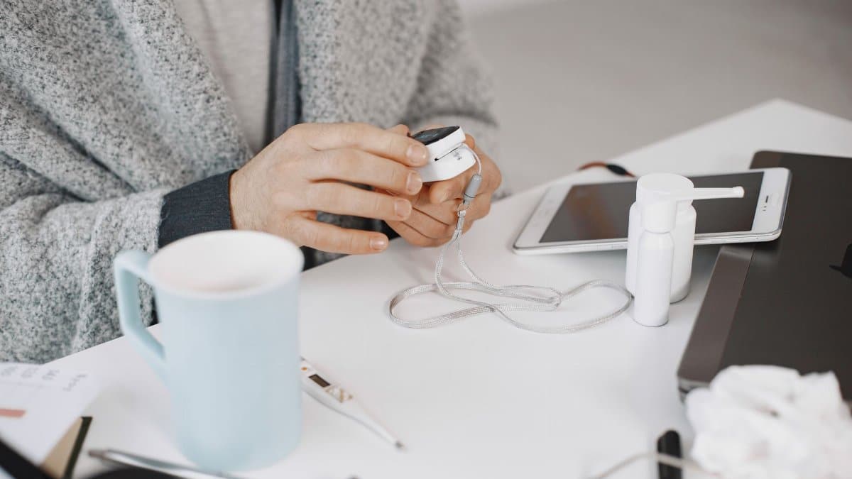 Close-up of hands using a health device at a desk with electronic gadgets and a cup.