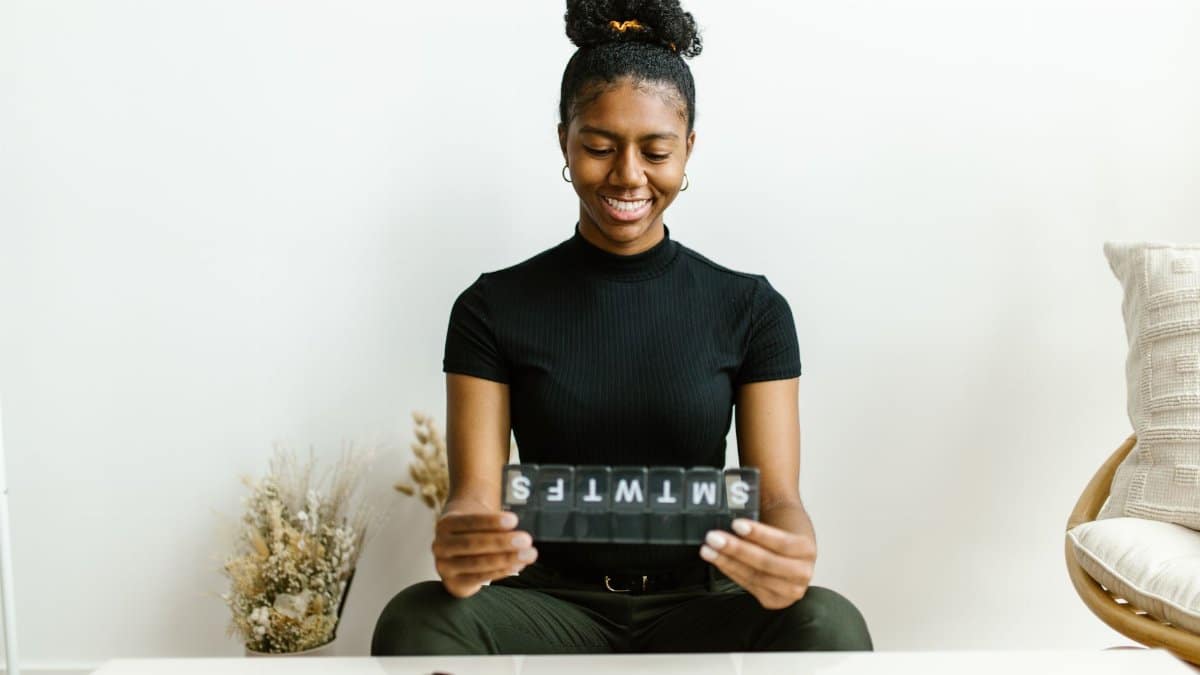 Black woman smiling while holding a weekly pill container, promoting health awareness.