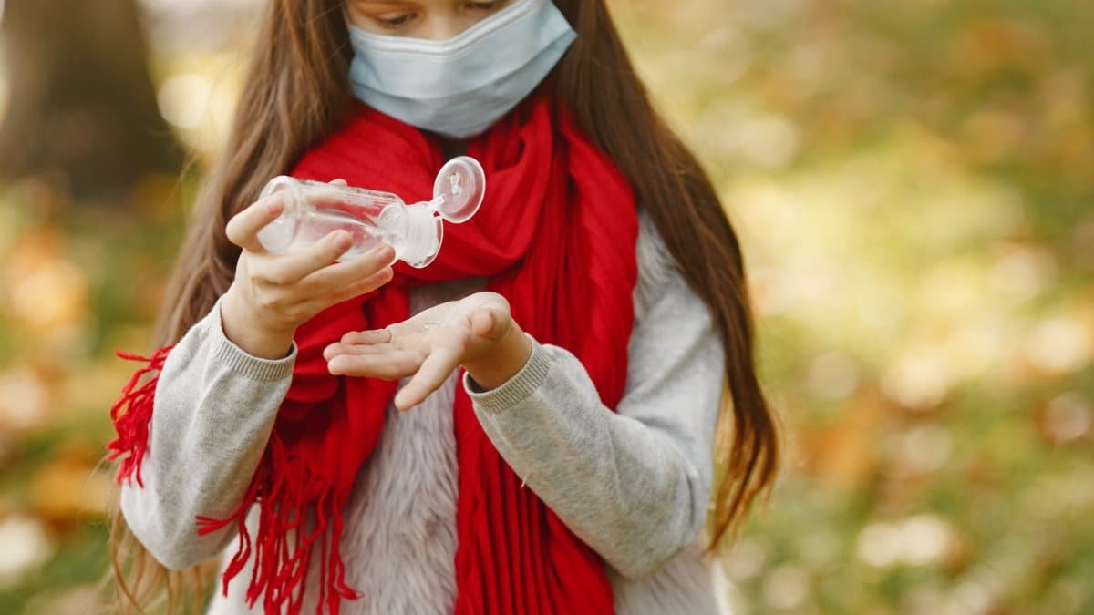 A young girl wearing a mask and red scarf applies hand sanitizer in a park during autumn.