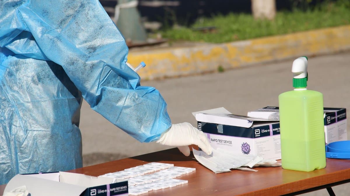 Healthcare worker in protective gear conducting rapid COVID-19 testing outdoors. PPE and test kits visible.
