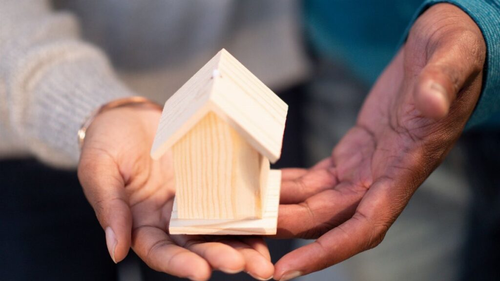 Hands holding a small wooden house model, symbolizing new home ownership and real estate transactions.