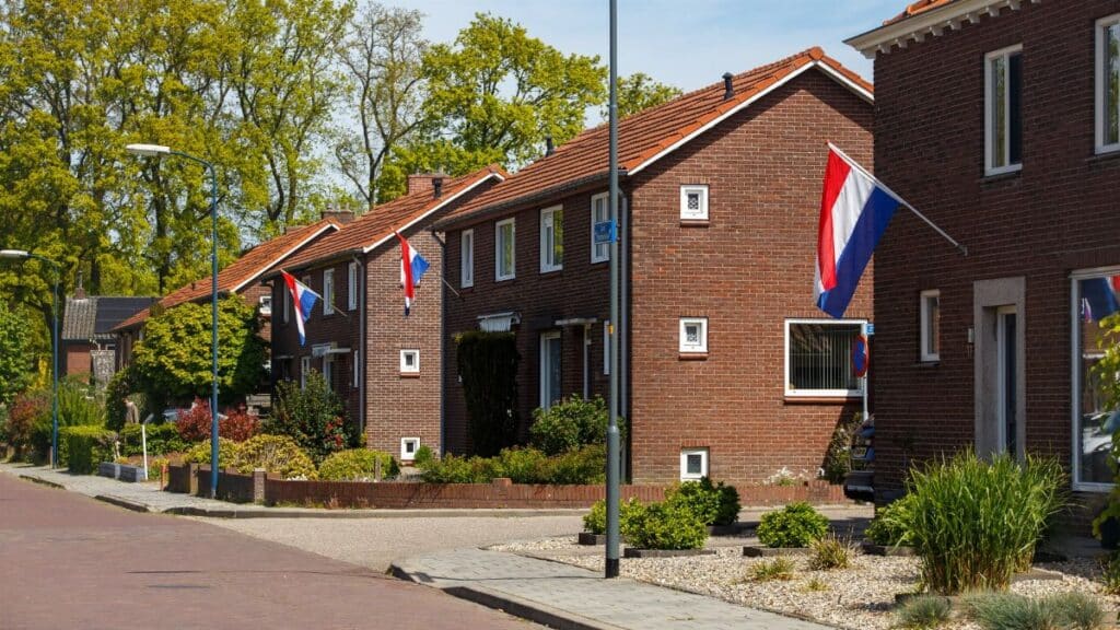 A picturesque suburban street in the Netherlands with Dutch flags displayed on brick houses during a sunny day.