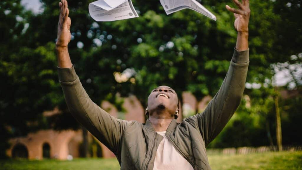 Happy African American remote worker tossing papers in air happy to get rid of boring paperwork while sitting in green park
