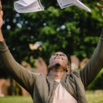 Happy African American remote worker tossing papers in air happy to get rid of boring paperwork while sitting in green park