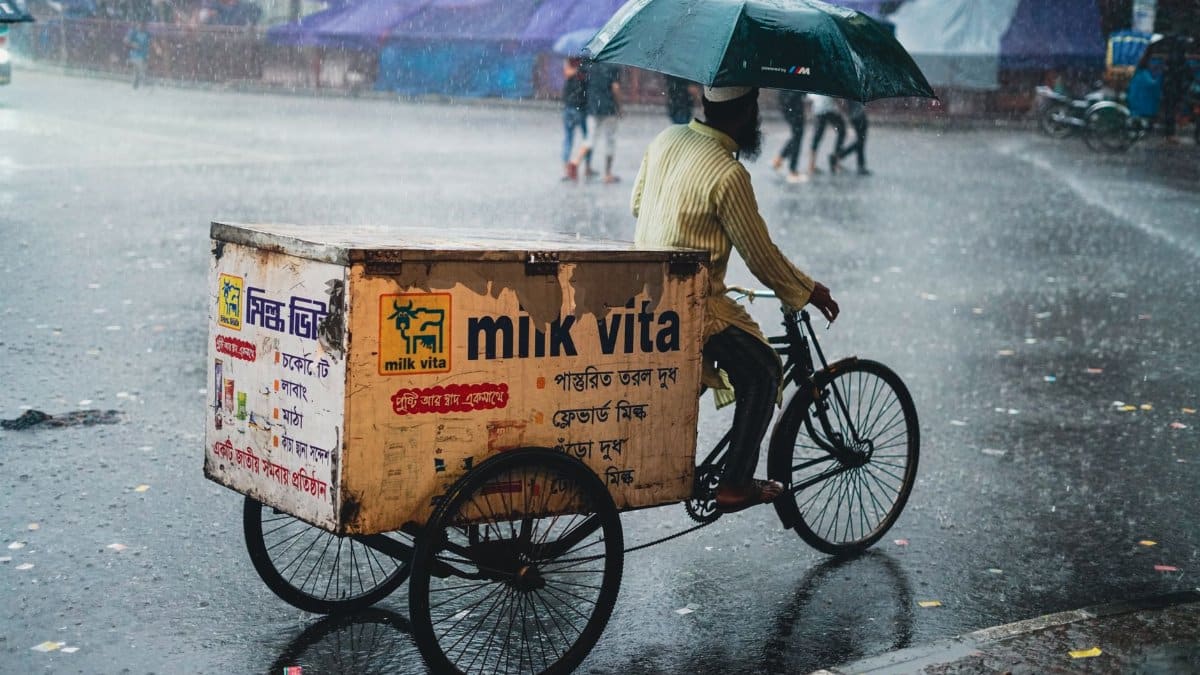 Man riding cycle rickshaw with umbrella during rain in Dhaka, street scene.