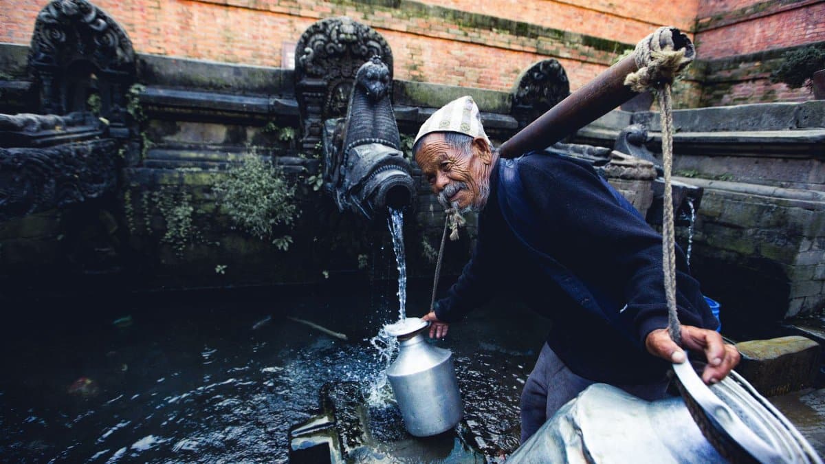 An elderly man gathers water from a historical stone fountain in Kathmandu, Nepal.