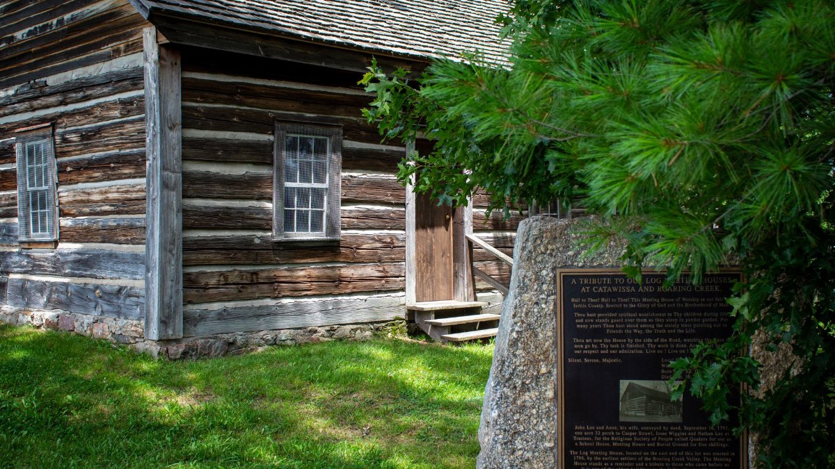 A rustic log cabin with a historical plaque surrounded by green foliage.
