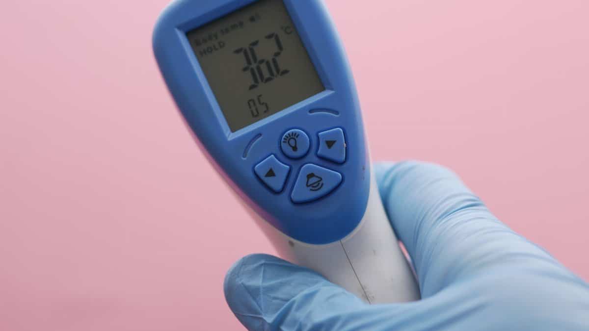 Close-up of an infrared thermometer displaying temperature, held by a gloved hand against a pink background.