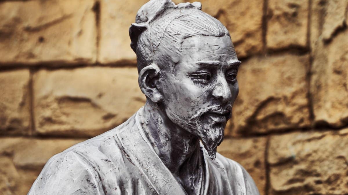 A detailed bronze sculpture of a historical figure set against a textured brick wall background.