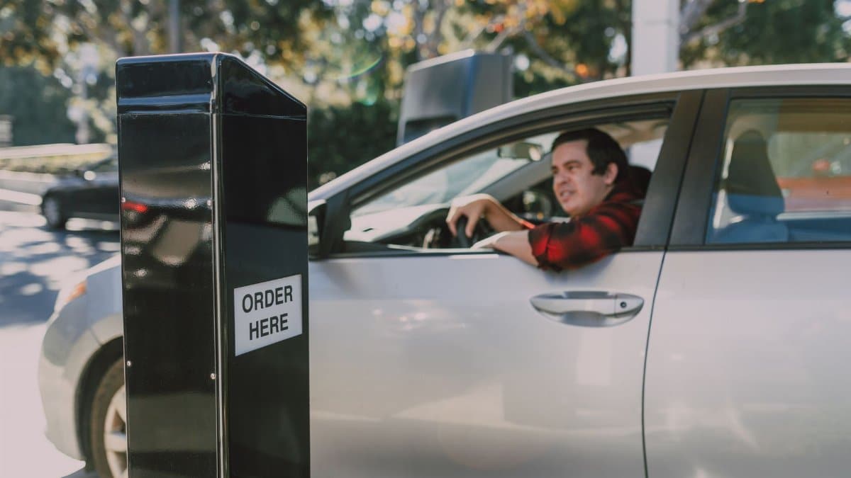 A man in a car orders at a drive-thru window, surrounded by outdoor scenery.