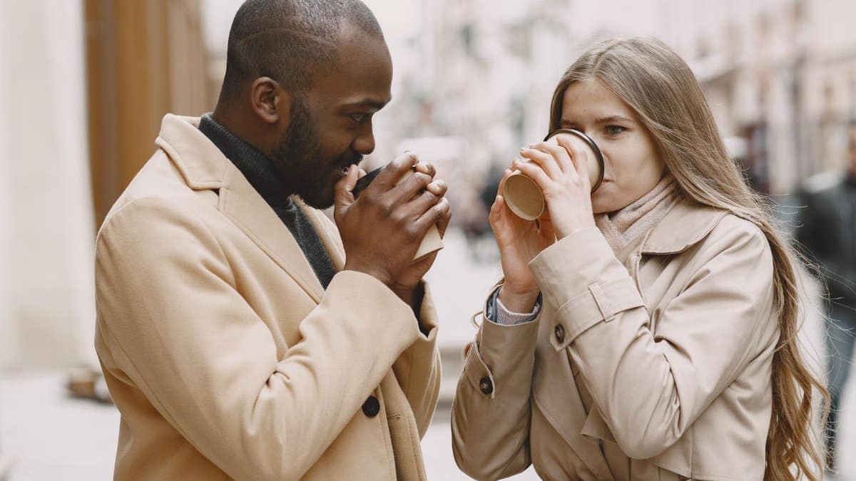 A stylish interracial couple drinks coffee outdoors in fall.