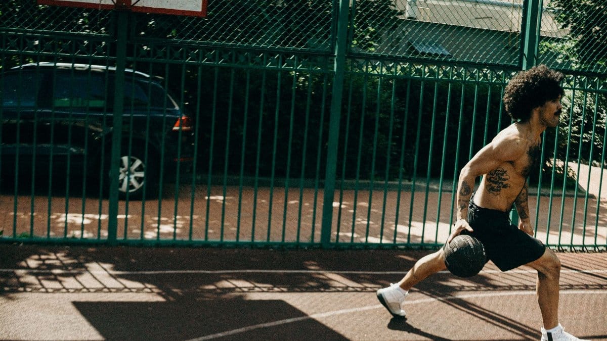 Shirtless man with tattoos performing fitness exercise with medicine ball on outdoor basketball court.
