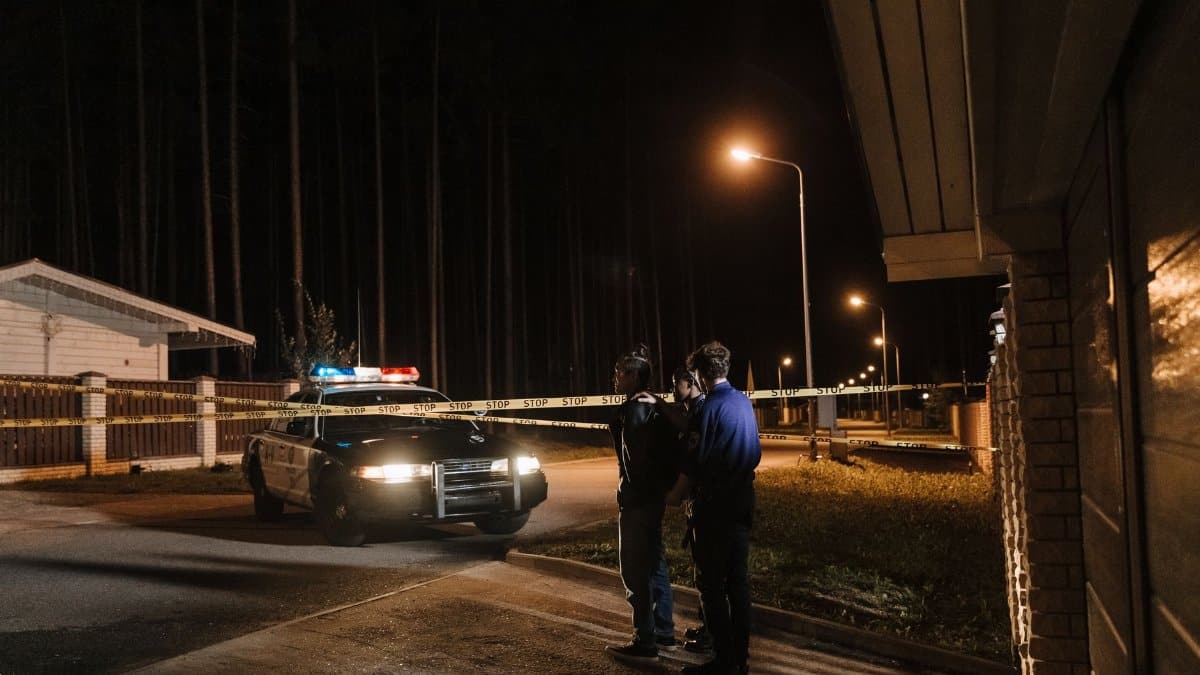 Police officers at a nighttime crime scene with a police car and caution tape under streetlights.