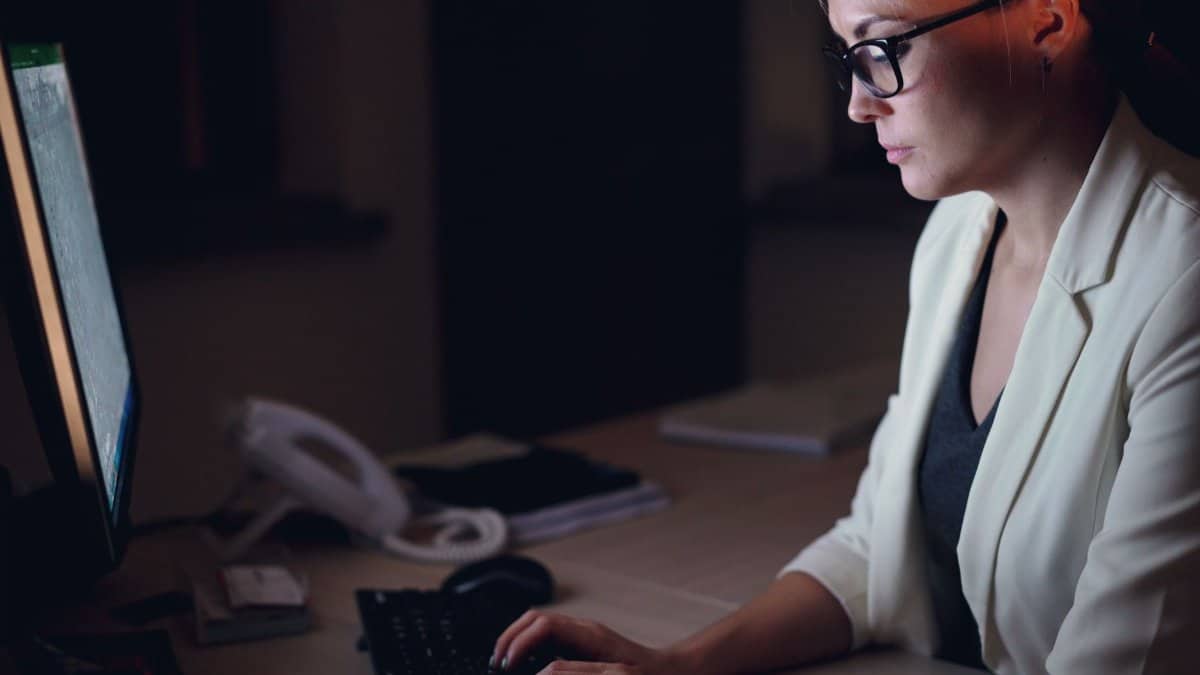 Woman in glasses working late at a desk with a computer monitor.