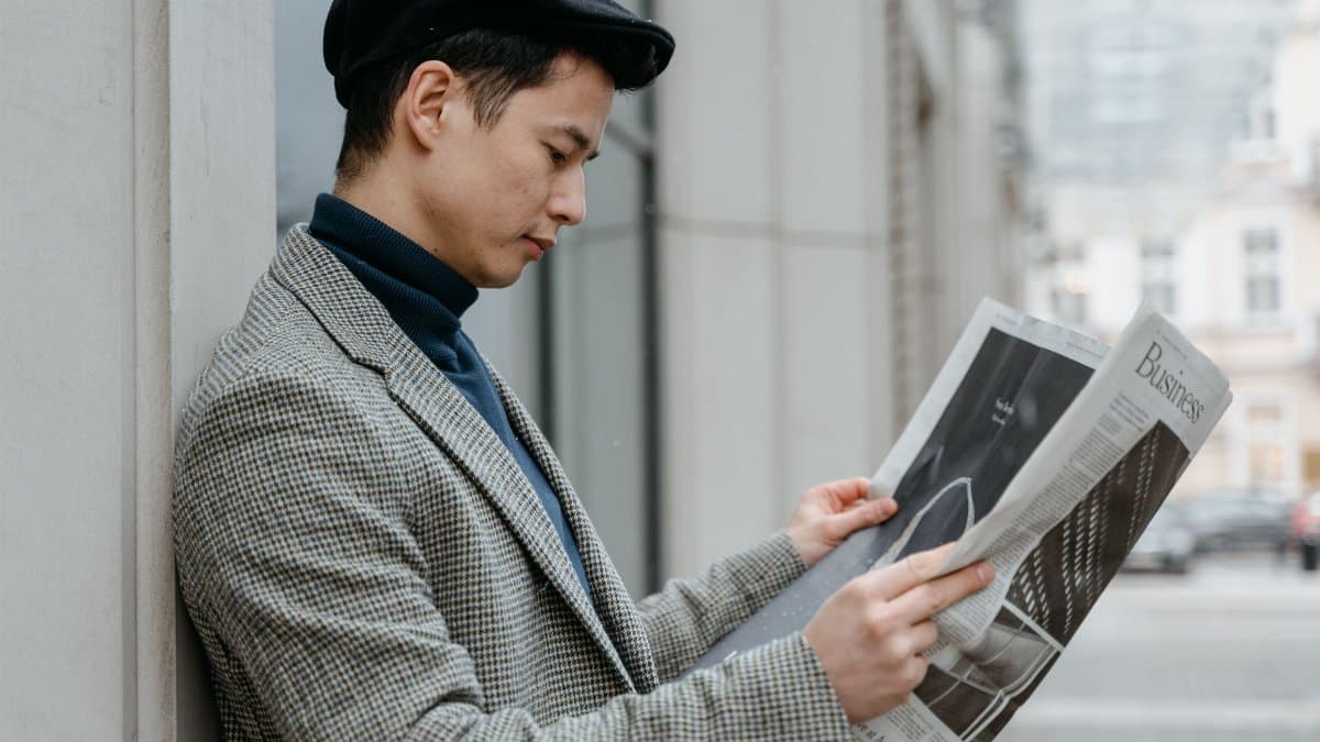 Side profile of a man reading a newspaper outdoors in an urban setting.