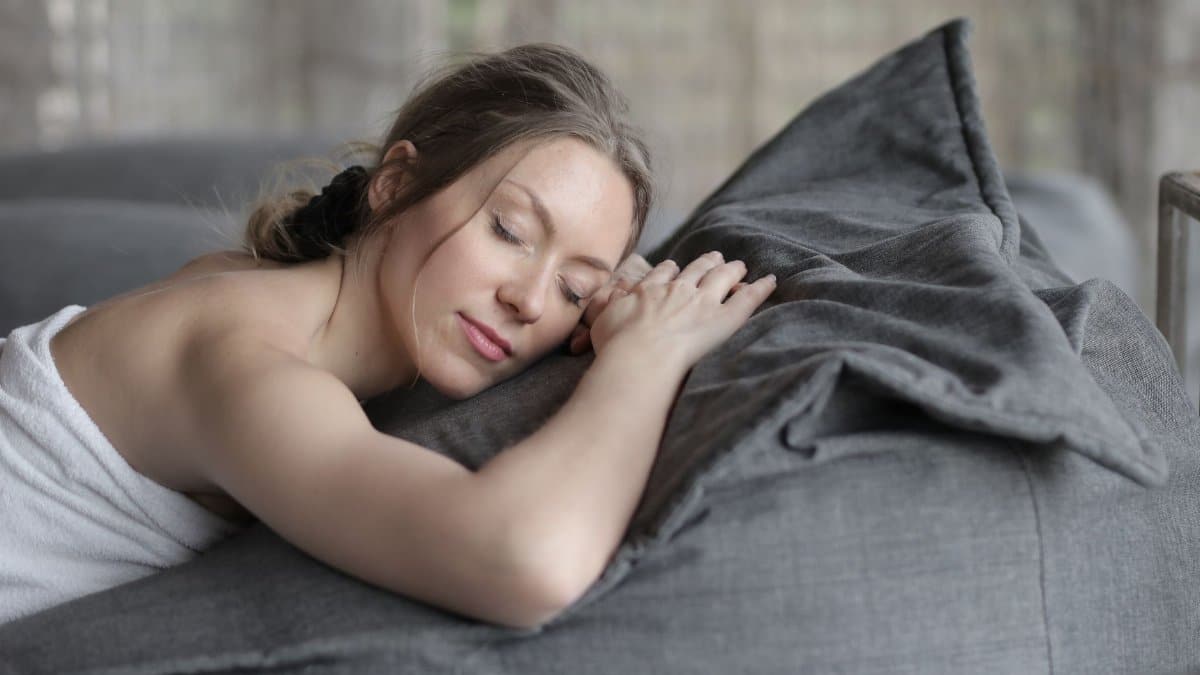 Peaceful woman with closed eyes sleeping on a cozy gray couch, wrapped in a towel indoors.