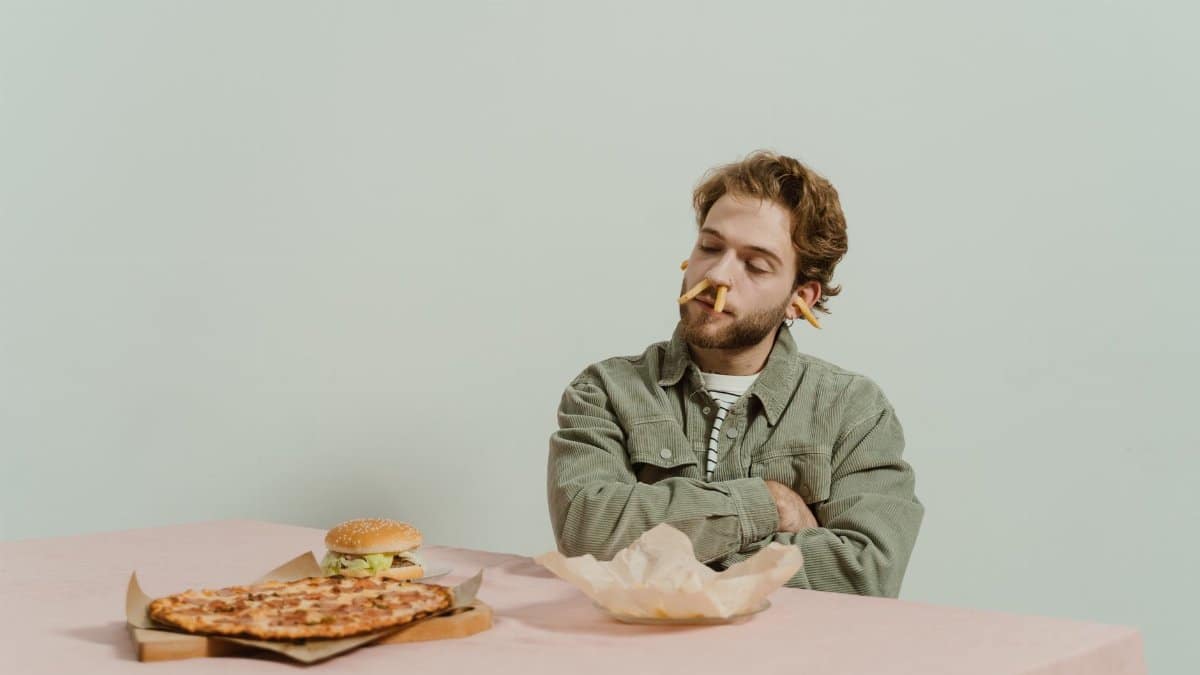 Bearded man with playful expression sitting at table with fast food, arms crossed. Conceptual indoors shoot.