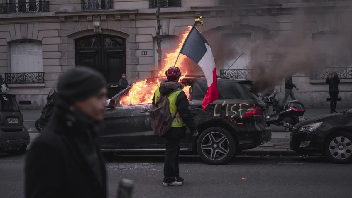Man in yellow vest holding French flag near burning car during protest in Paris.