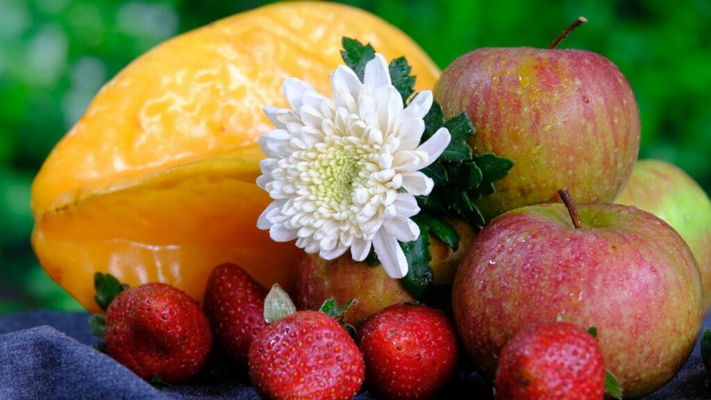 Vibrant still life of apples, strawberries, starfruit, and a chrysanthemum on a dark fabric backdrop.
