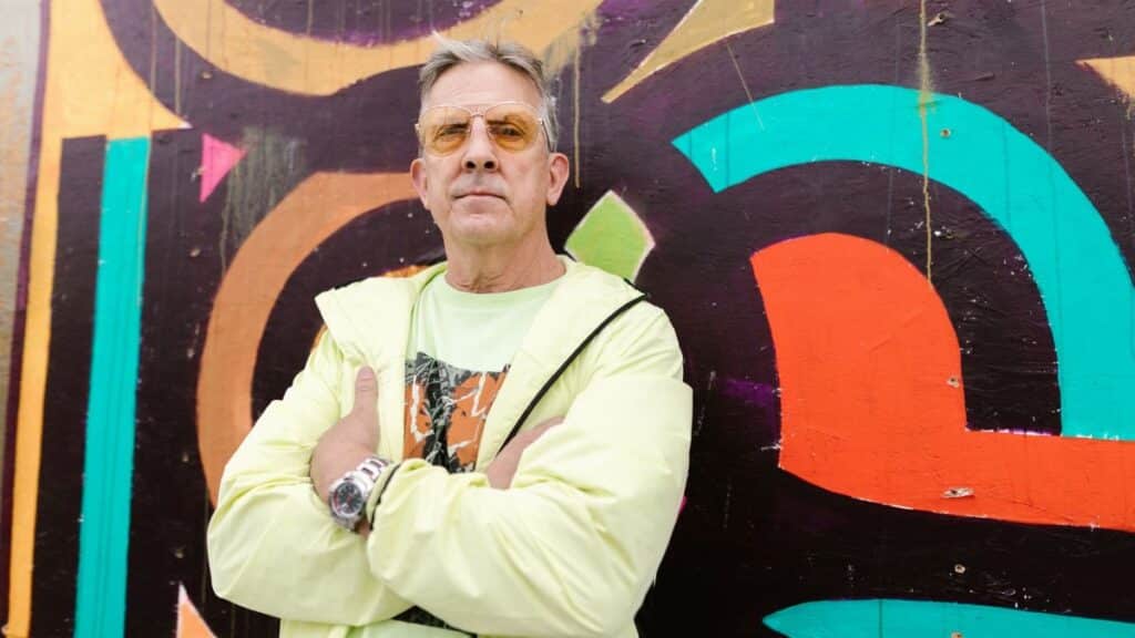 A confident senior man in bright attire poses against vibrant street art, exuding style.