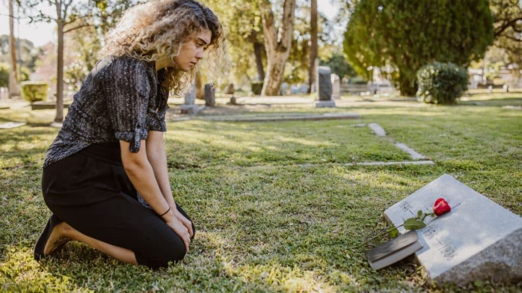 A woman kneeling by a grave in a sunlit cemetery, expressing grief and remembrance.