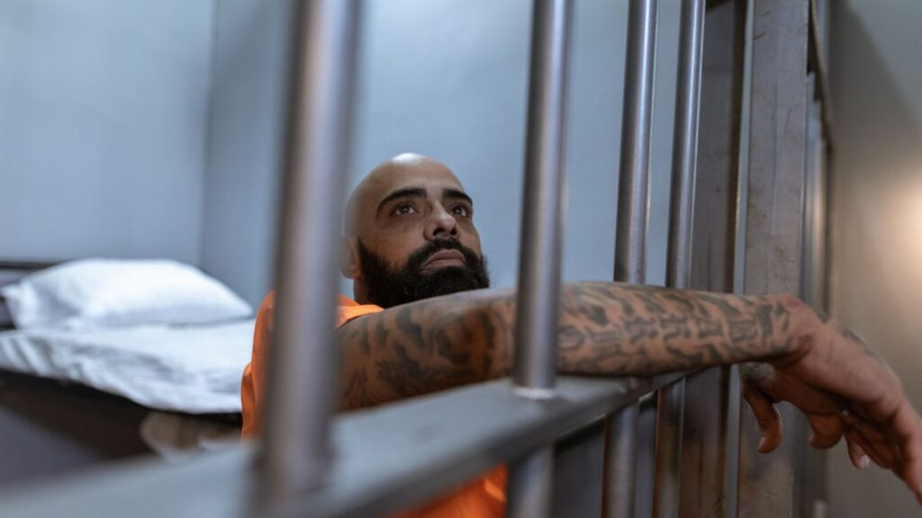 Tattooed inmate in an orange uniform behind prison bars, contemplating in a dimly lit cell.