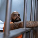 Tattooed inmate in an orange uniform behind prison bars, contemplating in a dimly lit cell.