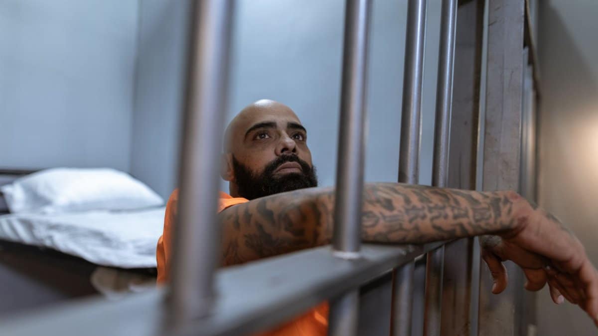 Tattooed inmate in an orange uniform behind prison bars, contemplating in a dimly lit cell.