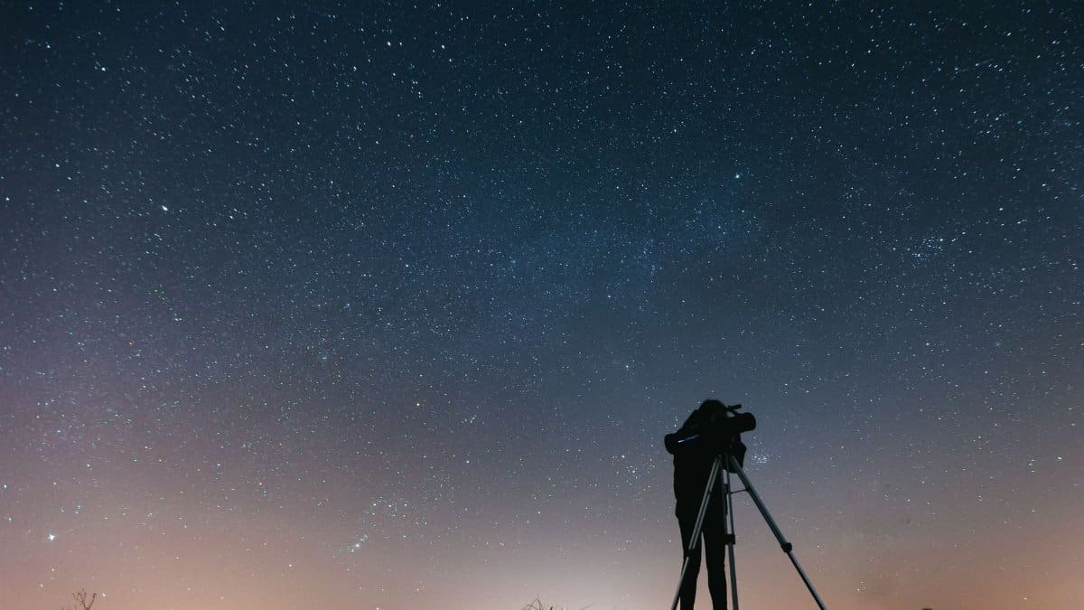 A silhouetted person using a telescope to observe the night sky filled with stars.