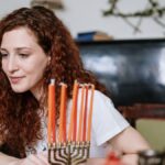 Woman with red hair sits by a menorah with lit candles, celebrating a Jewish holiday indoors.