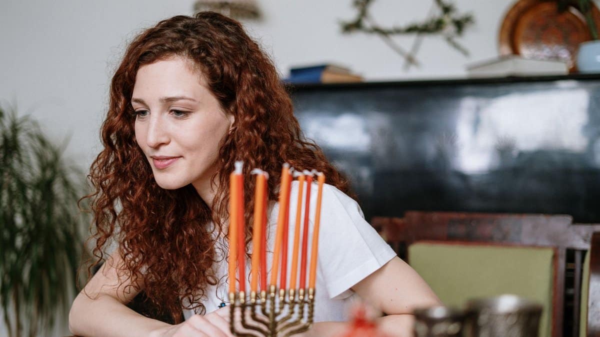 Woman with red hair sits by a menorah with lit candles, celebrating a Jewish holiday indoors.