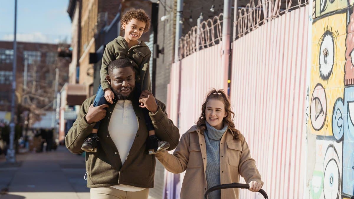 Positive African American father with son on shoulders and happy mother with baby stroller strolling on sunny street in city