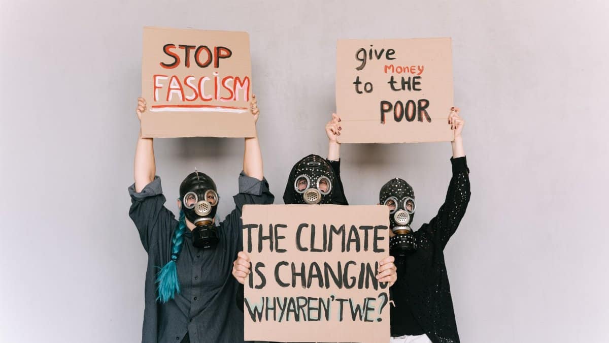 Three people wearing gas masks hold signs advocating social change and awareness.