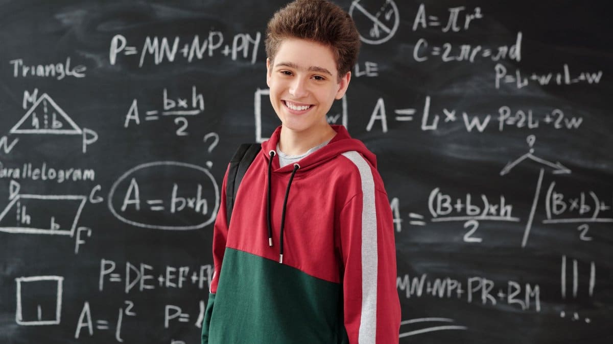 Smiling student in a classroom posing in front of a chalkboard filled with math formulas.