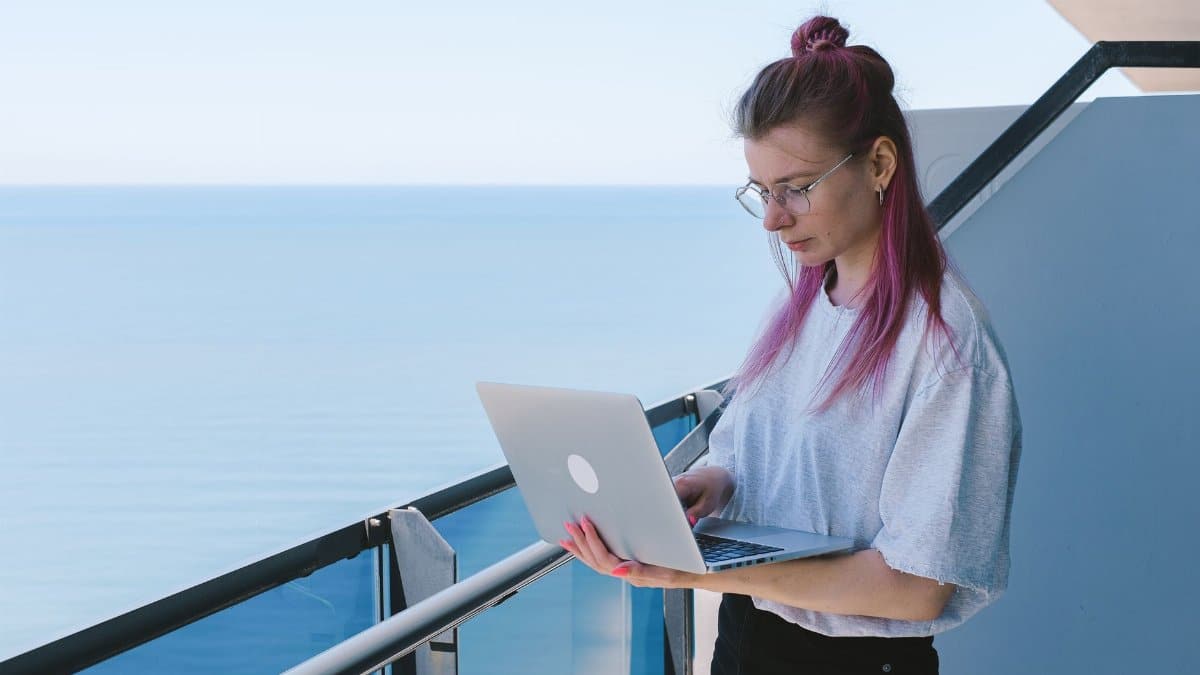 A young woman works remotely on her laptop by the sea on a sunny day, embodying the digital nomad lifestyle.