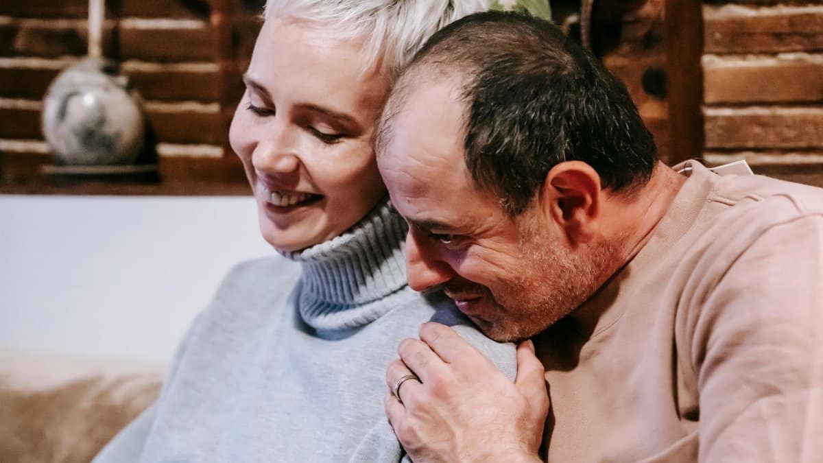 Side view of enamored middle aged male in casual clothes hugging smiling wife while resting together on comfortable sofa at home