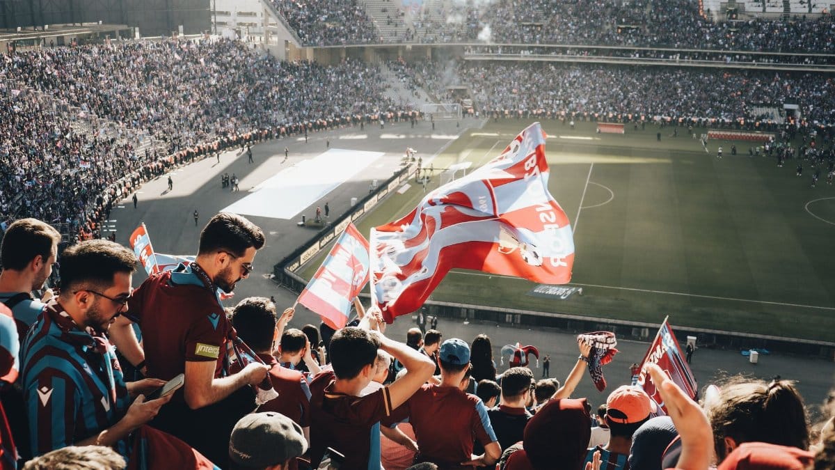 Vibrant crowd with flags cheering at a football stadium during the day.