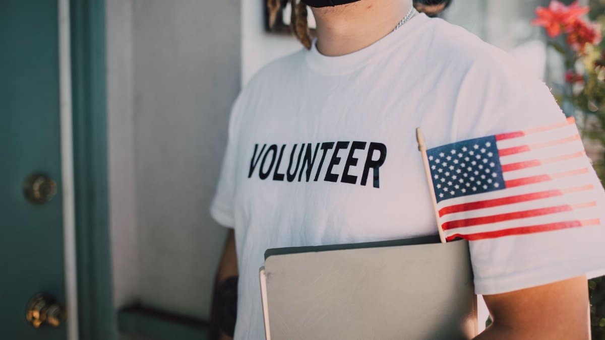 Volunteer wearing a mask holds a notebook and American flag, symbolizing community service.