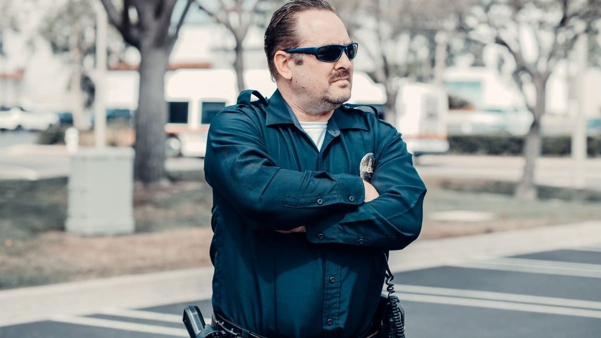 A police officer in uniform stands confidently outdoors with arms crossed and sunglasses.
