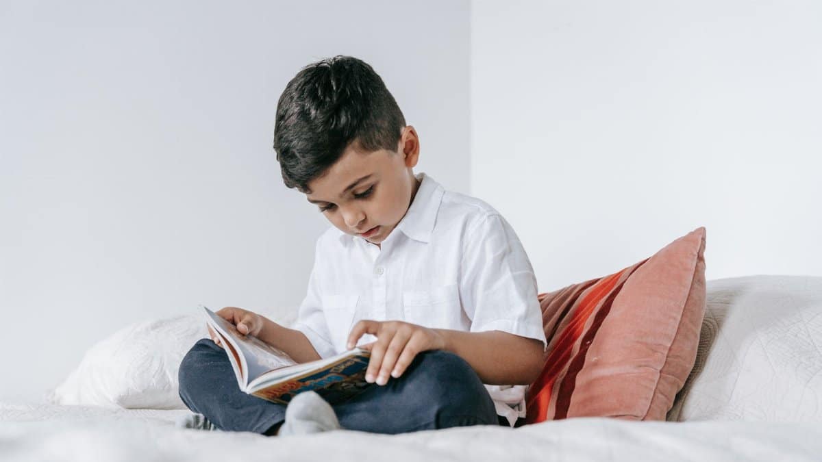 A young boy intently reading a book while sitting on a bed at home.