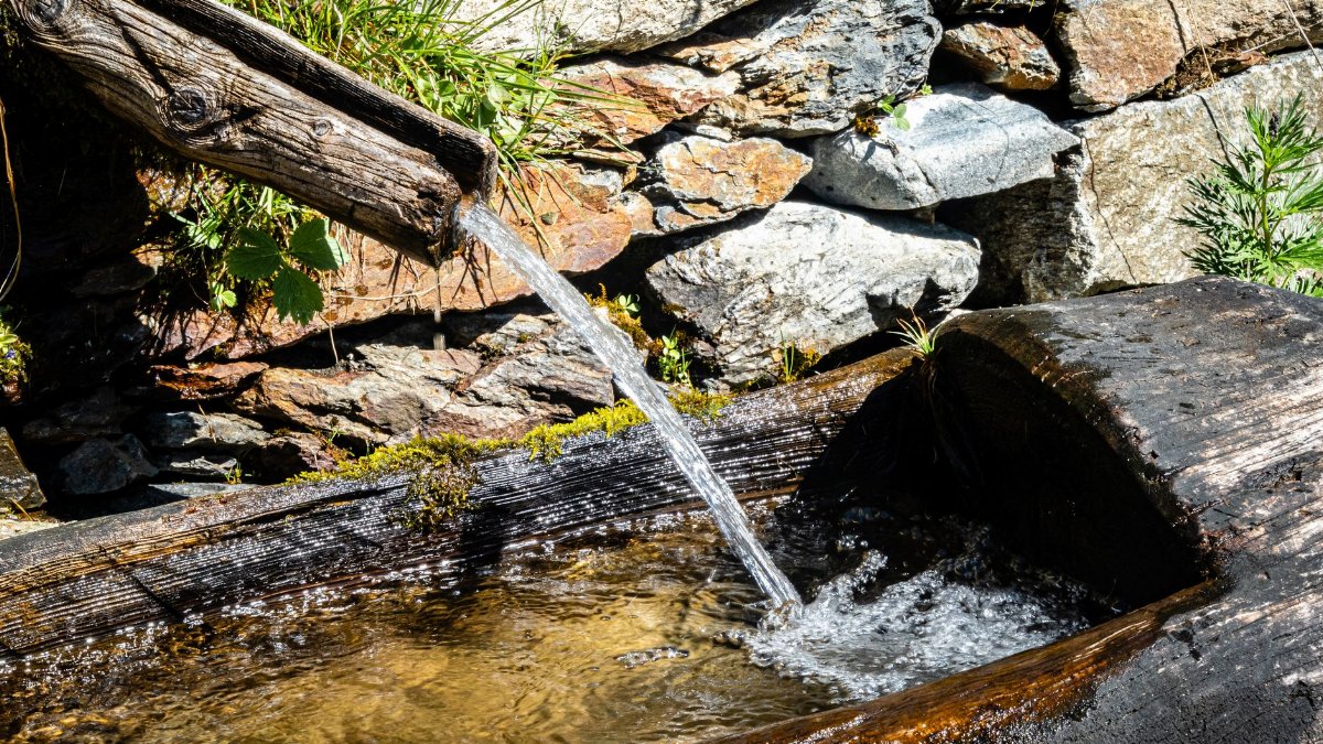 Clear water flows from a rustic wooden spout into a basin, set against a stone wall.