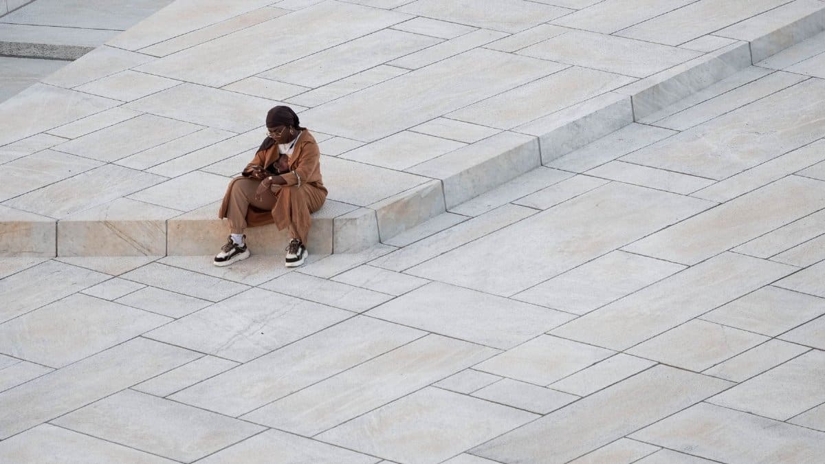 A woman sits alone on geometric paved steps in an urban environment, using her smartphone.